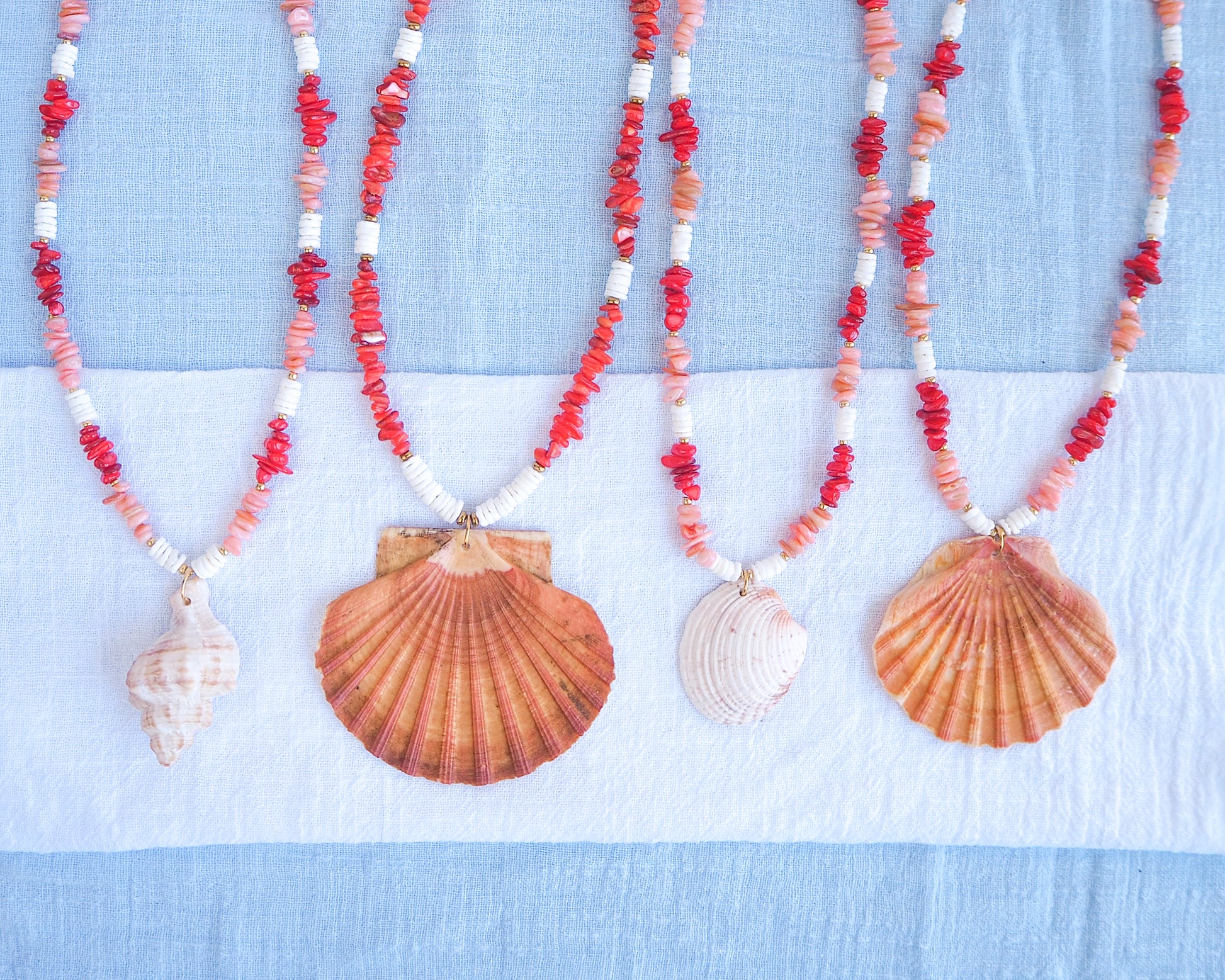 Necklaces with shell pendants and red coral stone beads on a blue and white background. A collection of big shell pendant necklaces and red and white beads on a light blue background. Big scallop shell necklace paired with red stone beads, pink stone beads and white shell beads. The shell is a great scallop shell from Portugal and is about 7 cm. The color of the shell is orange pink and it has vertical ridges. 