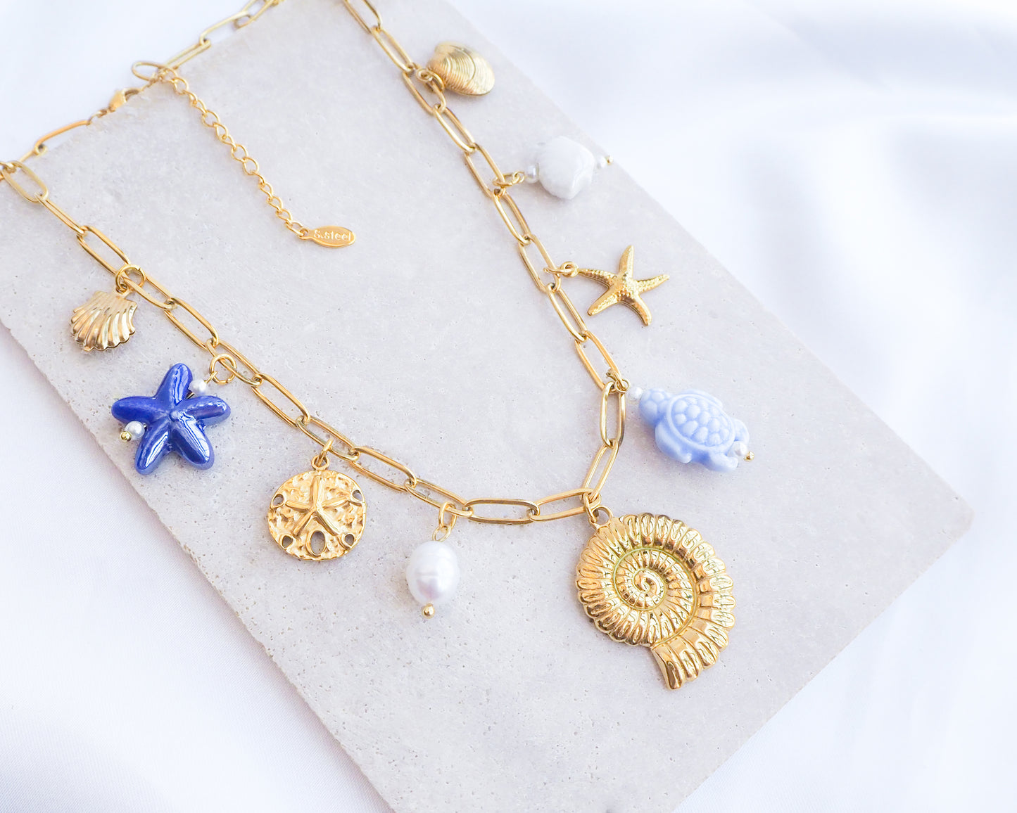 Blue Ocean Ceramic Charm Necklace with 3 blue ceramic charms (turtle, starfish, shell), Shell pendant, sand dollar pendant, starfish pendant, 18k gold stainless steel chain displayed on stone 