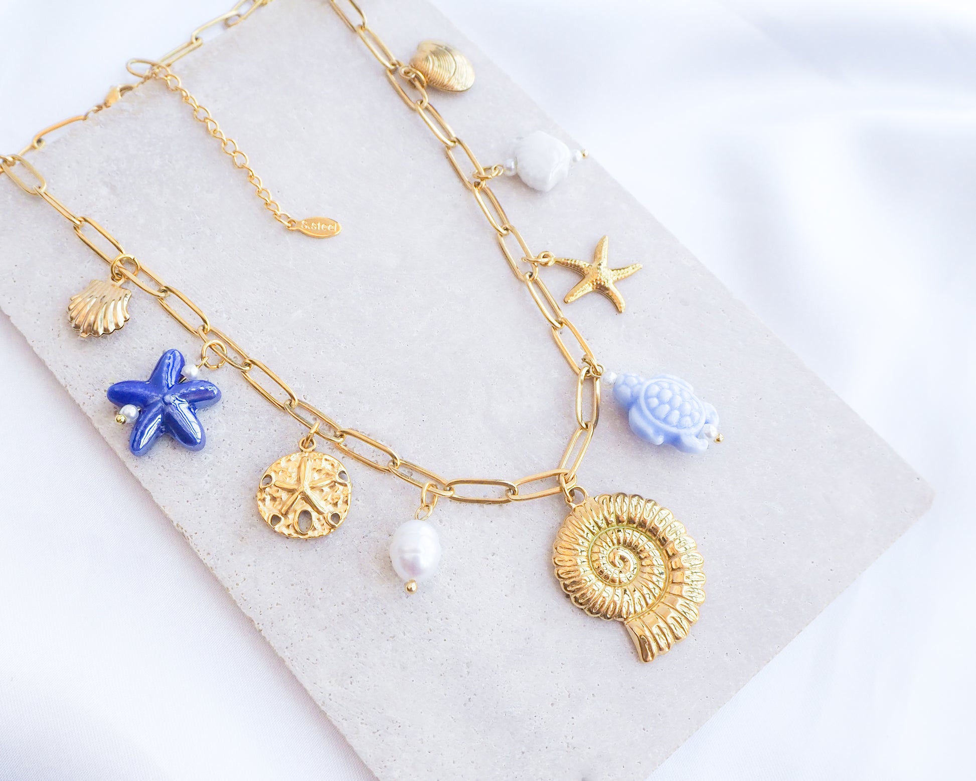 Blue Ocean Ceramic Charm Necklace with 3 blue ceramic charms (turtle, starfish, shell), Shell pendant, sand dollar pendant, starfish pendant, 18k gold stainless steel chain displayed on stone 