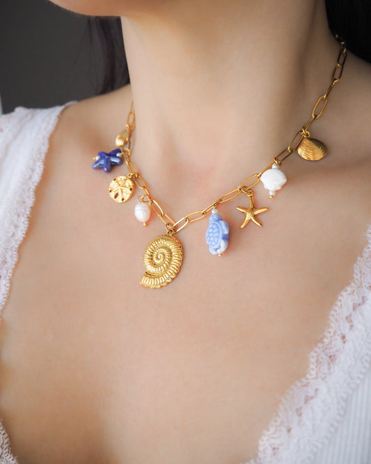 Model wearing Blue Ocean Ceramic Charm Necklace with 3 blue ceramic charms (turtle, starfish, shell), Shell pendant, sand dollar pendant, starfish pendant, 18k gold stainless steel chain displayed on stone 
