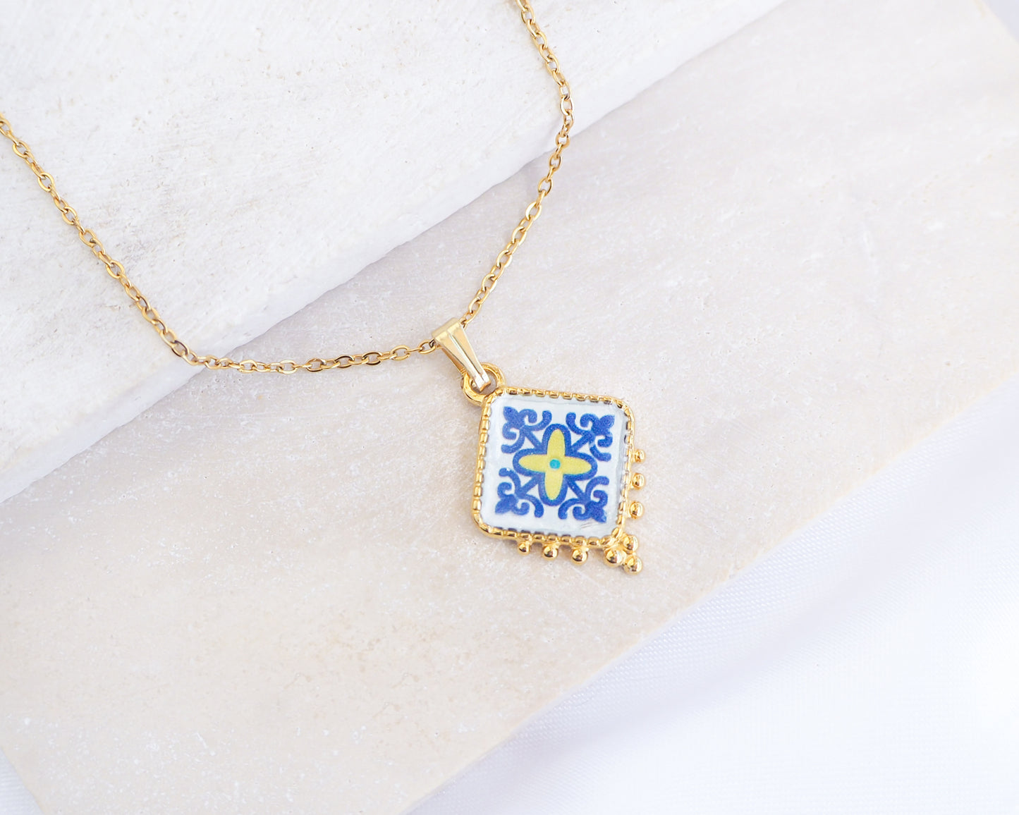 Close up of Blue Yellow Portuguese Tile Necklace Gold on stone displayed. Made with 18k gold stainless steel and blue yellow purple pendant inspired by the Portuguese Azulejo design pattern. 