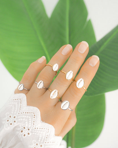 Hand of model wearing handmade rings with tiny cowrie shells from Portugal, European cowrie shell rings made with silver stainless steel and 18k gold stainless steel, adjustable beach jewelry from Portugal, hand is wearing a white sleeve and has a green plant background