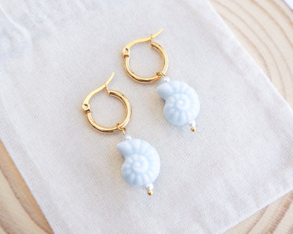 Gold ceramic seashell hoop earrings with light blue pherical charms on a white cloth background
