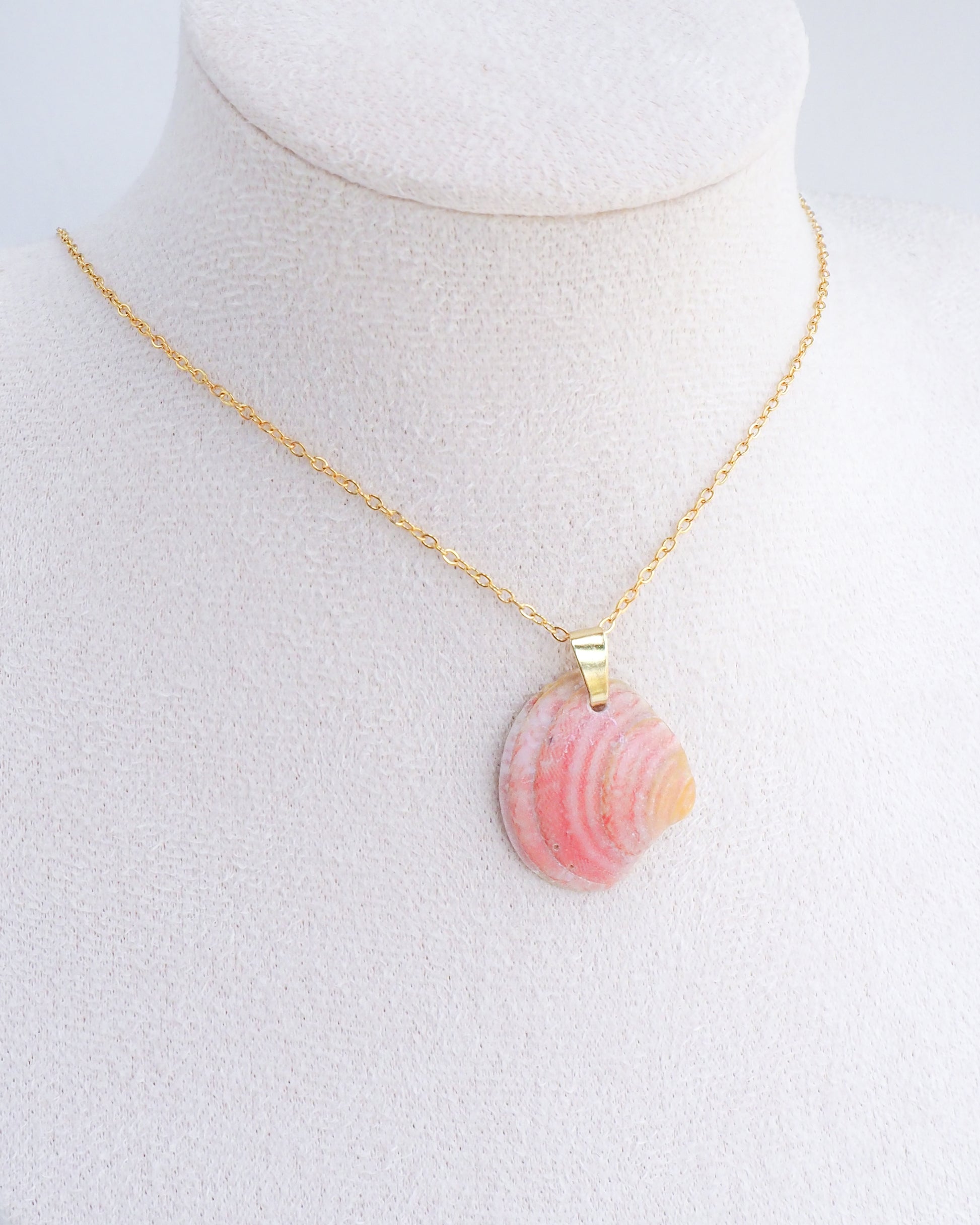 A neck display with gold chain necklace with a natural coral peach pink seashell pendant. The seashell is a venus shell handpicked from Portugal set in a gold clasp and gold necklace chain. Made of 18k gold stainless steel.