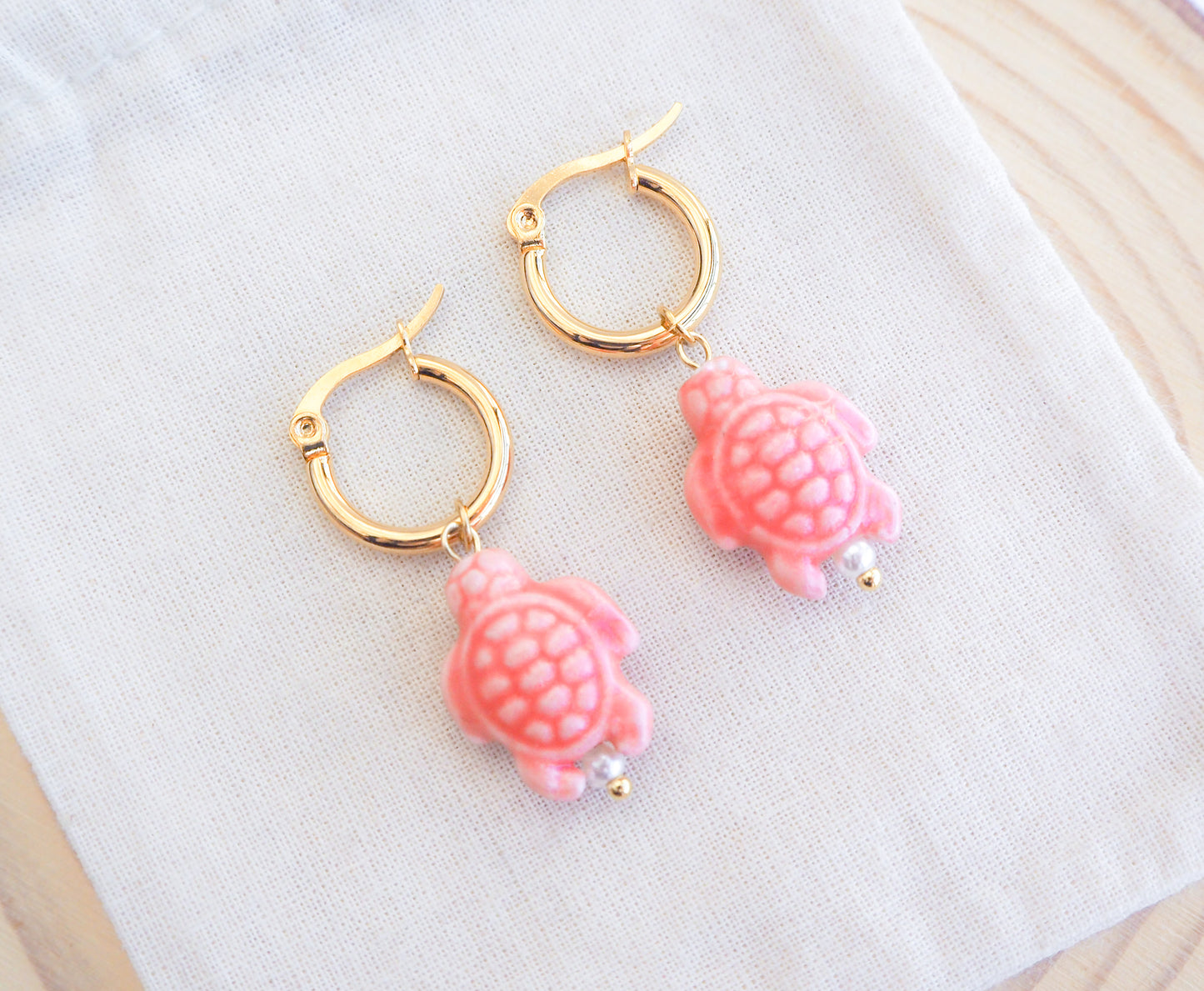 Gold hoop earrings with pink turtle ceramic charms on a white surface