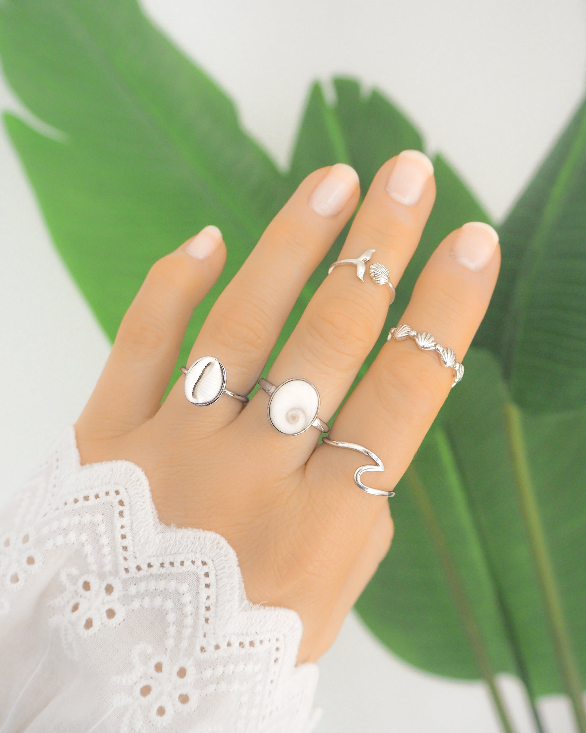 Hand wearing multiple silver rings with a green leaf background. Hand with silver tiny cowrie shell ring, a real shiva Eye of Saint Lucia silver ring, a whale tail ring silver, a silver wave ring and a silver horizontal shell ring, all adjustable, green plant background, tiny european cowrie shell from Portugal