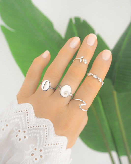 Hand wearing multiple silver rings with a green leaf background. Hand with silver tiny cowrie shell ring, a real shiva Eye of Saint Lucia silver ring, a whale tail ring silver, a silver wave ring and a silver horizontal shell ring, all adjustable, green plant background, tiny european cowrie shell from Portugal