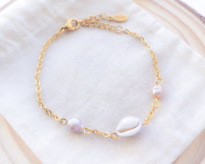 Close-up of cowrie shell pearl bracelet on cotton displayed. Handpicked European cowrie shell from Portugal set on a 18k gold stainless steel bracelet chain and adorned with 2 freshwater peals that are pink-purple in color. The shell is about 8 mm and white in color. 