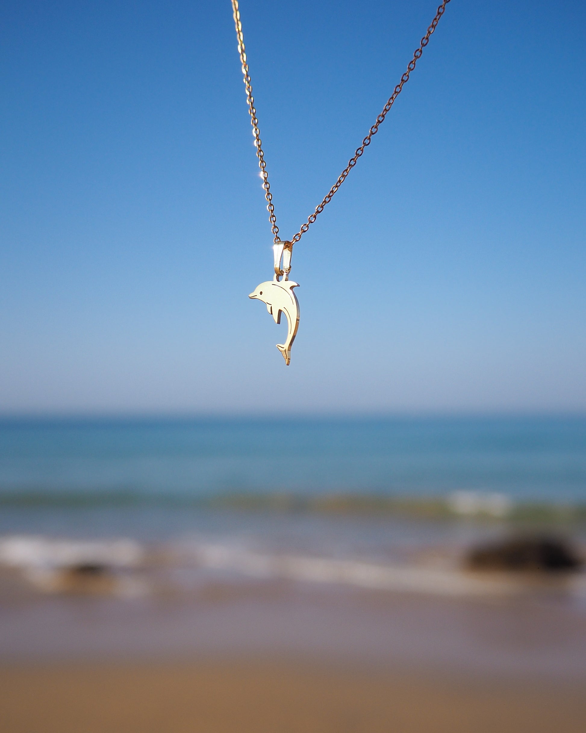 Dolphin Minimalistic Gold Necklace a chain against a beach backdrop with blue sky and ocean.