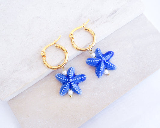 Dark Blue Ceramic Sea Star Gold Hoop Earrings on stone displayed, 14k gold stainless steel earring hoops with blue ceramic starfish pendants, ocean inspired jewelry from Portugal