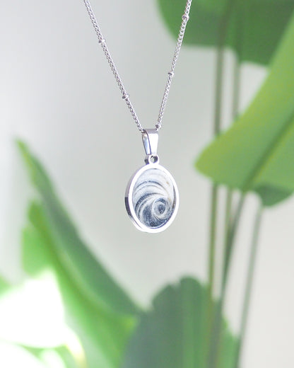 Front view of Dark Grey Eye of Saint Lucia Silver Necklace with green plant background, silver stainless steel shiva eye pendant necklace, Saint Lucy Eye, operculum pendant