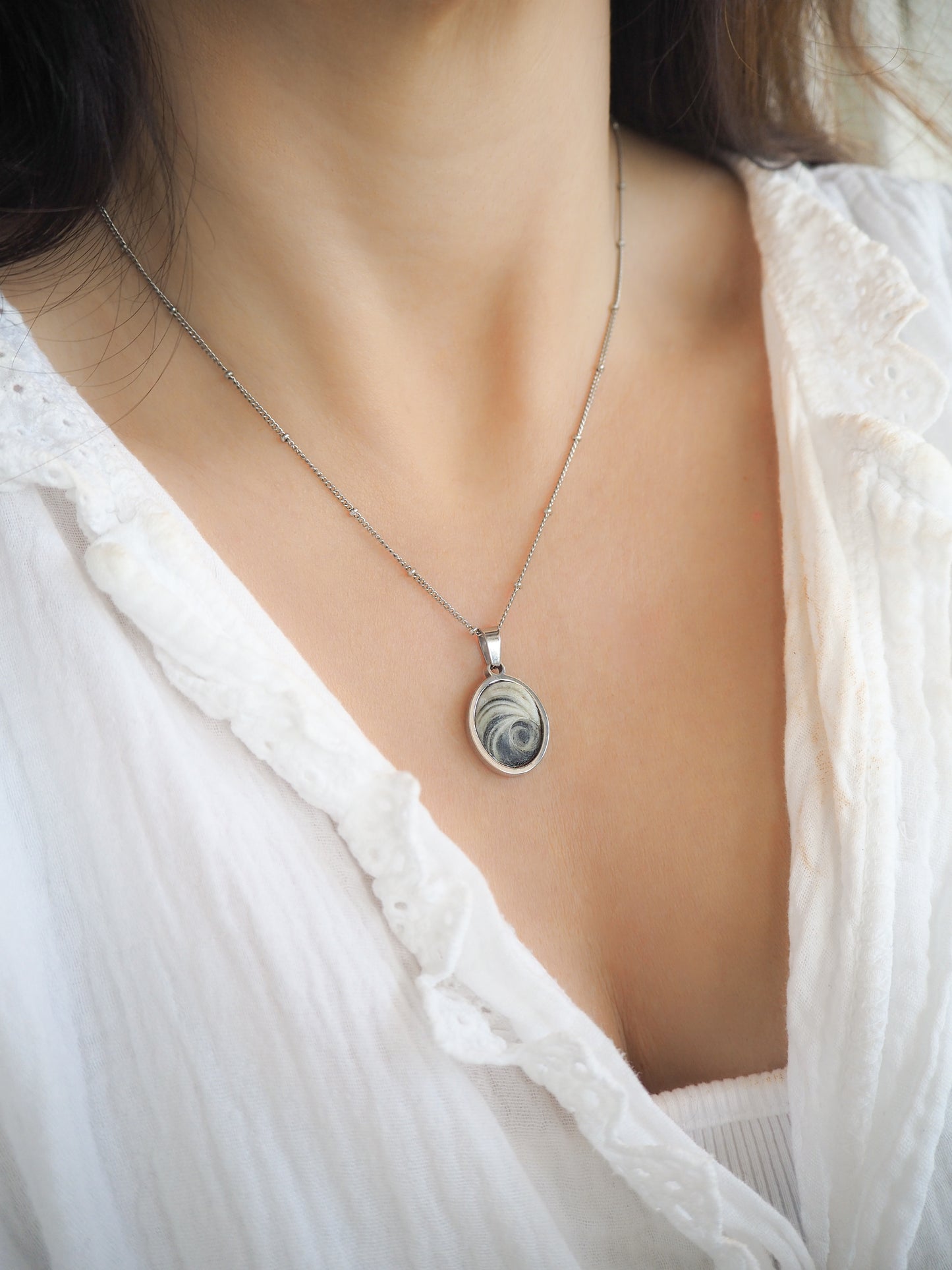 Model wearing Dark Grey Eye of Saint Lucia Silver Necklace on neck with white blouse and brown hair, silver stainless steel shiva eye pendant necklace, Saint Lucy Eye, operculum pendant