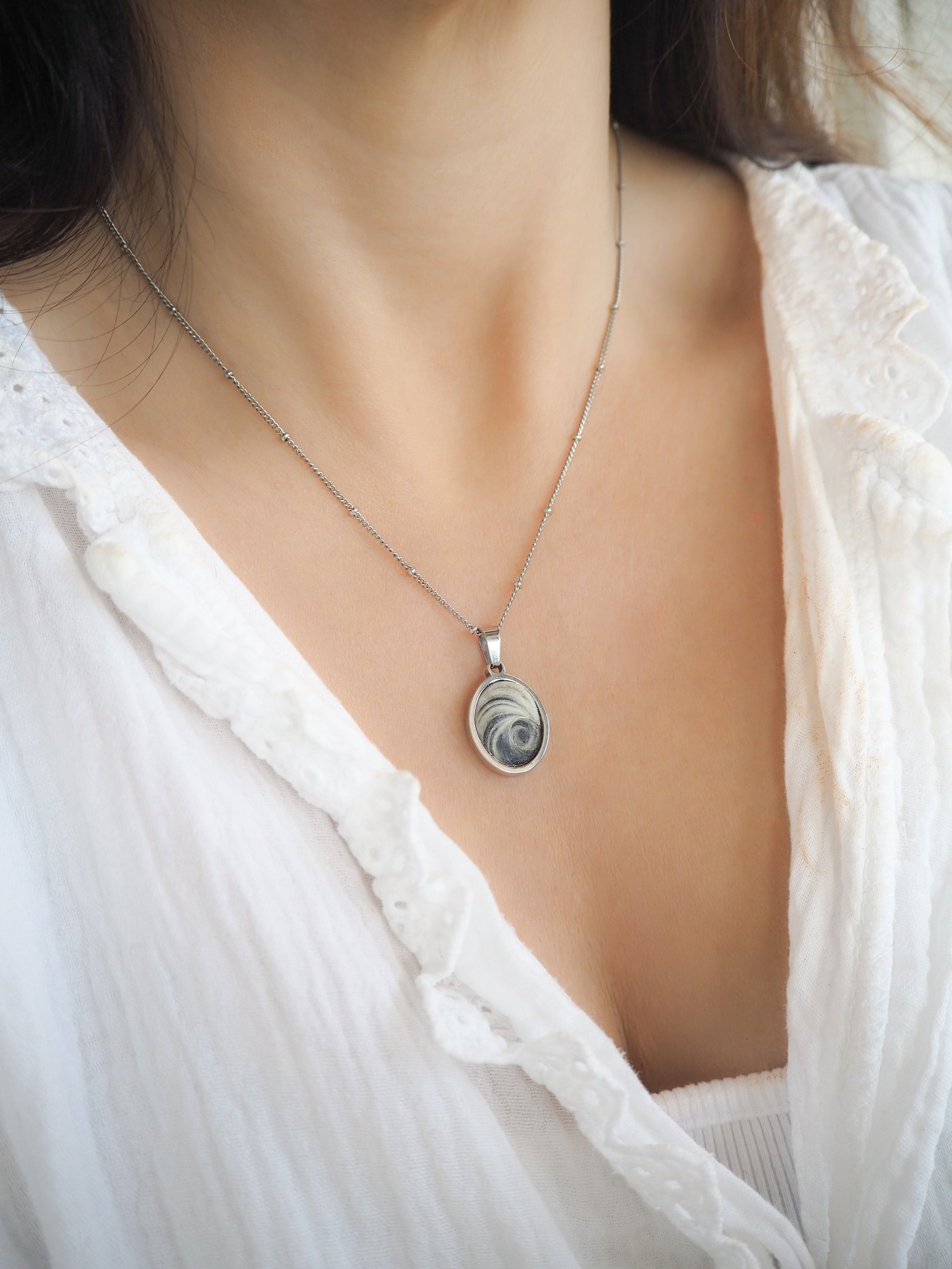 Model wearing Dark Grey Eye of Saint Lucia Silver Necklace on neck with white blouse and brown hair, silver stainless steel shiva eye pendant necklace, Saint Lucy Eye, operculum pendant