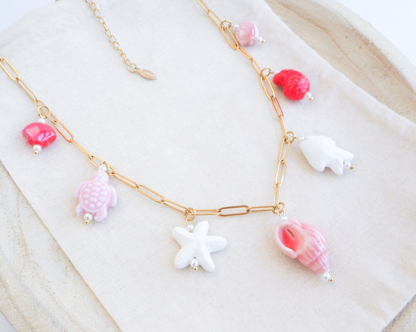 Close up of Sweet Ocean Ceramic Charm Necklace with 7 ceramic ocean charms (turtle, starfish, fish, shell & more), Pink white red sea pendants, 18k gold stainless steel chain displayed on light background