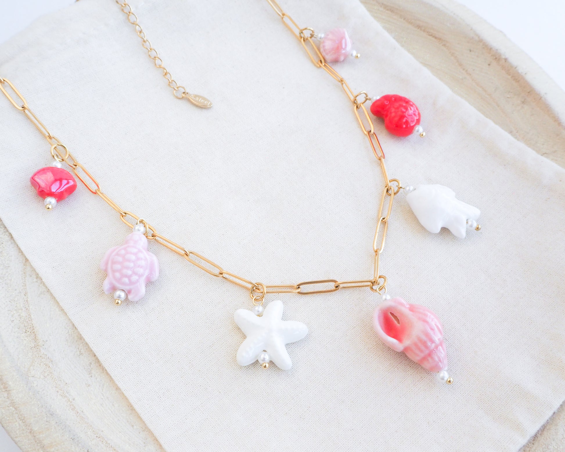 Close up of Sweet Ocean Ceramic Charm Necklace with 7 ceramic ocean charms (turtle, starfish, fish, shell & more), Pink white red sea pendants, 18k gold stainless steel chain displayed on light background
