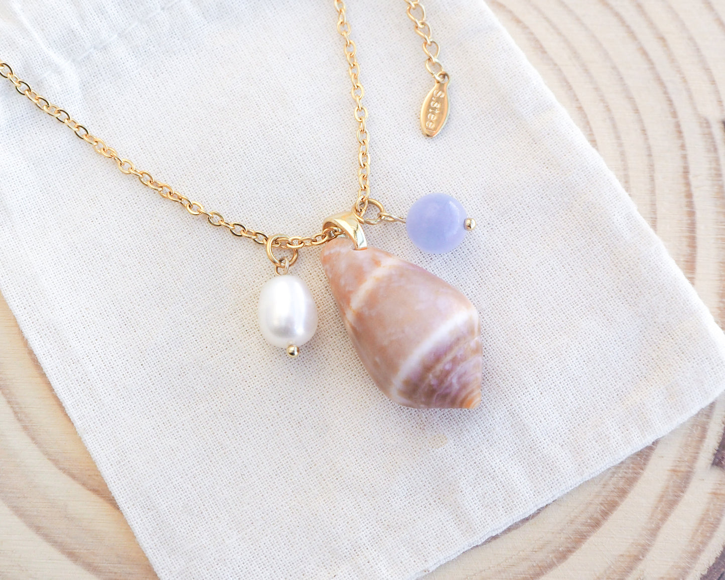 Close up of gold necklace with real cone shell and a gemstone and pearl necklace on display. Mediterranean cone shell was handpicked from Portugal. Made with a ivory white freshwater pearl and blue angelite gemstone bead. Made with 18k gold stainless steel clasp and necklace chain. 