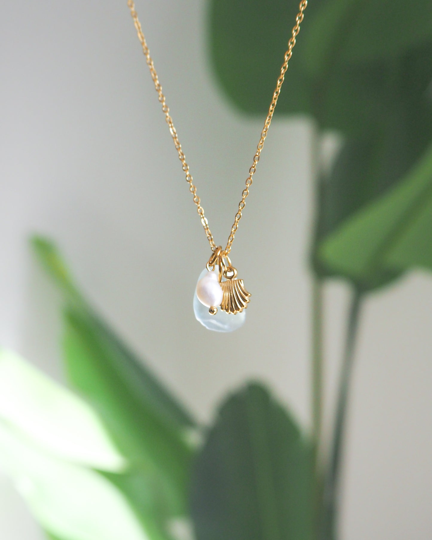 Front view of Oceanic Trio Shell Light Blue Sea Glass Pearl Necklace with green plant background, scallop shell necklace, ocean blue necklace, coastal jewelry, freshwater pearl necklace, gold seashell necklace, ocean-inspired jewelry, beach necklace, ocean treasure necklace