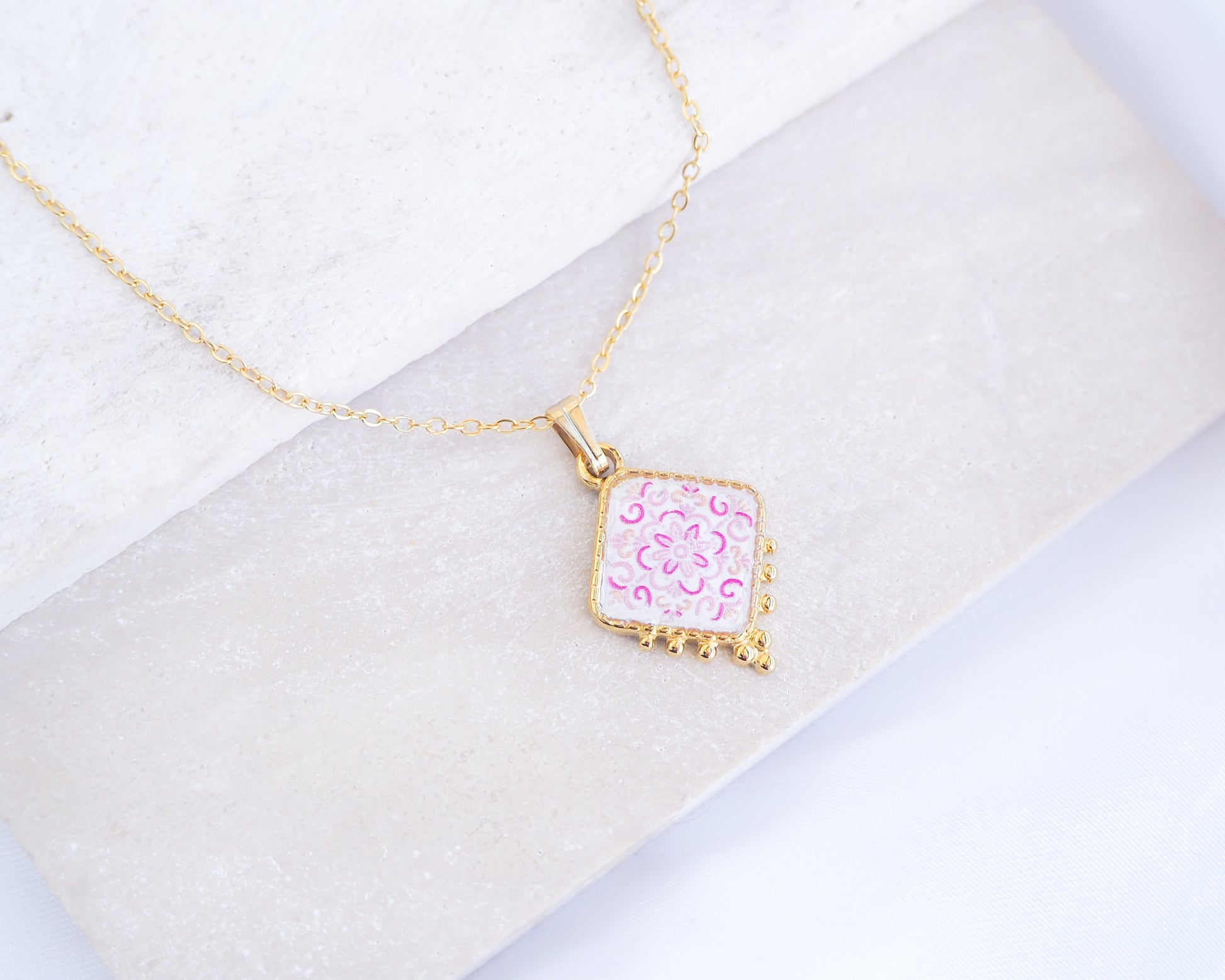 Close up of Portuguese Tile Necklace Floral Pink Gold on stone displayed. Made with 18k gold stainless steel and white pink floral pendant inspired by the Portuguese Azulejo design pattern. 