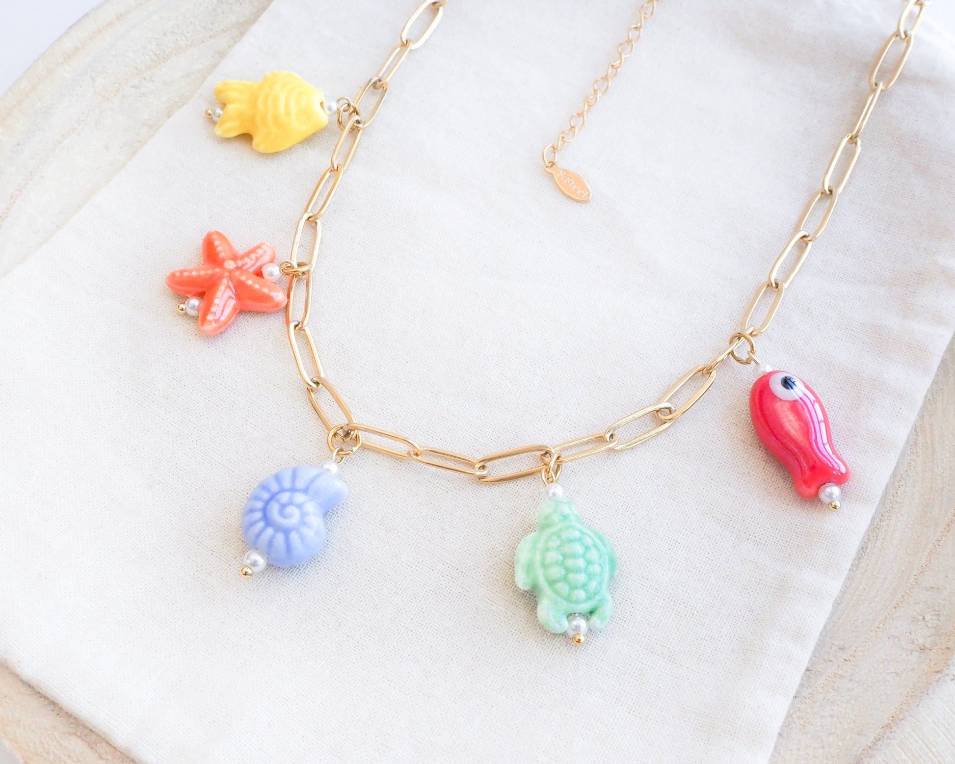 Side view of Rainbow Ceramic Charm Necklace with 5 ceramic ocean charms (turtle, starfish, fish, shell & more), sea treasure pendants, 18k gold stainless steel chain displayed on stone 