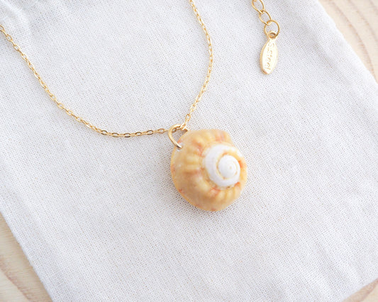 Side view of Sunny Magus Top Shell Necklace on display. A gold chain necklace with a delicate orange seashell pendant. The seashell is a orange in colour with a white interior. It's a handpicked magus top shell from the beach of Portugal.