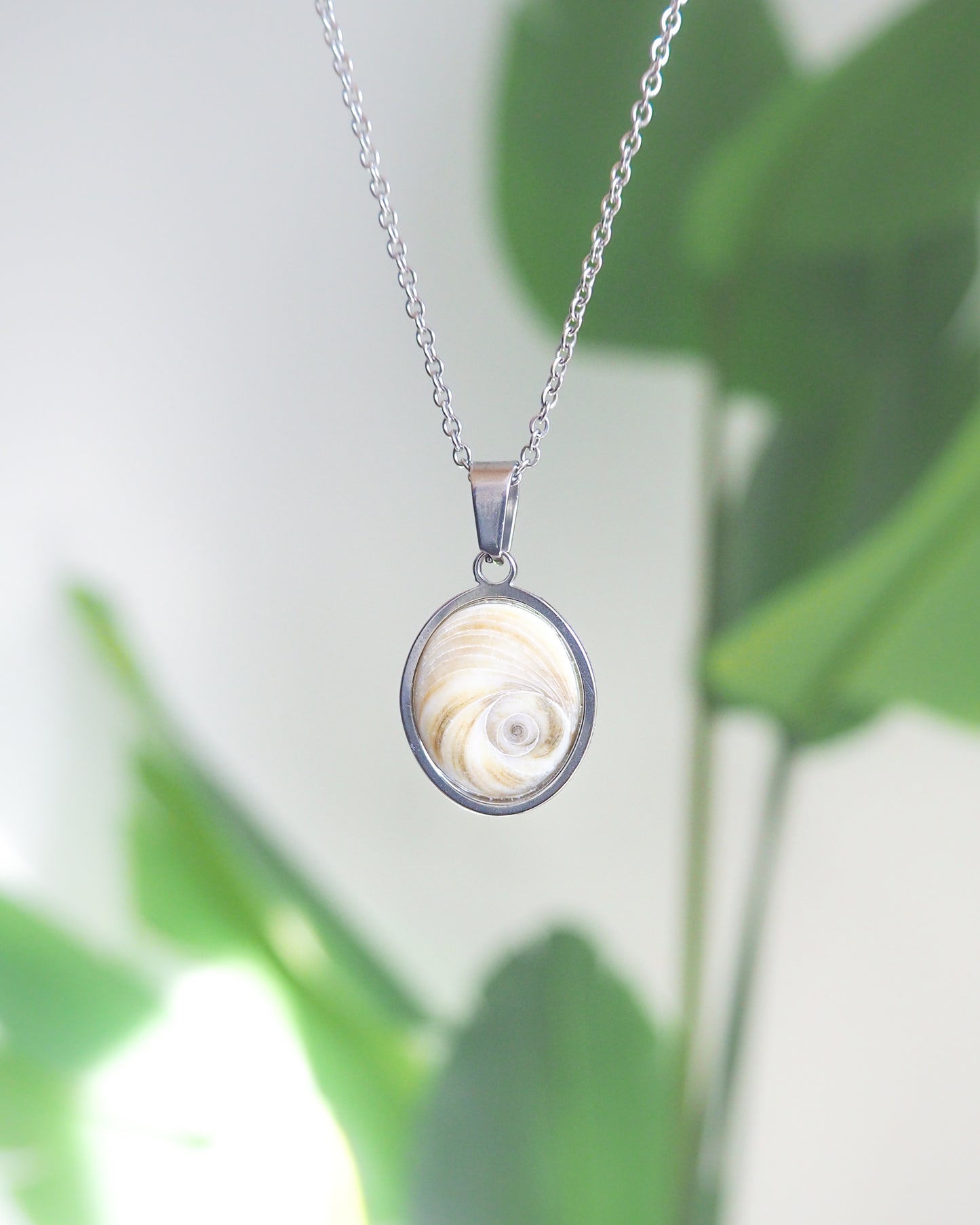 Front view of Light Eye of Saint Lucia Silver Necklace with green plant background, silver stainless steel shiva eye pendant necklace, Saint Lucy Eye, operculum pendant