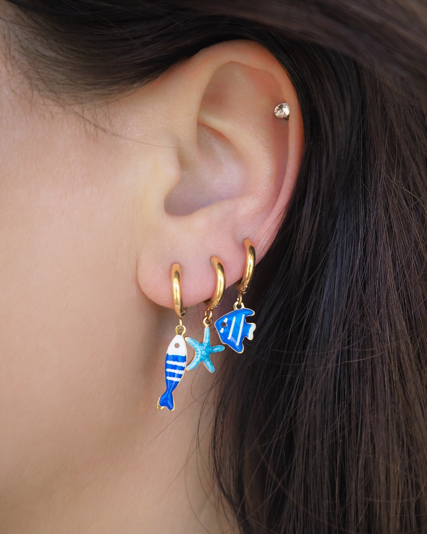 Model wearing a tiny blue sardine fish, tiny starfish and blue fish hoop earring in ear. The starfish pendants are blue. Made with 18k gold stainless steel earring hoops. Beach jewelry for summer days. Minimalistic coastal earrings. Perfect to mix and match with other earrings.