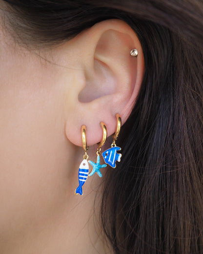 Model wearing a tiny blue sardine fish, tiny starfish and blue fish hoop earring in ear. The starfish pendants are blue. Made with 18k gold stainless steel earring hoops. Beach jewelry for summer days. Minimalistic coastal earrings. Perfect to mix and match with other earrings.