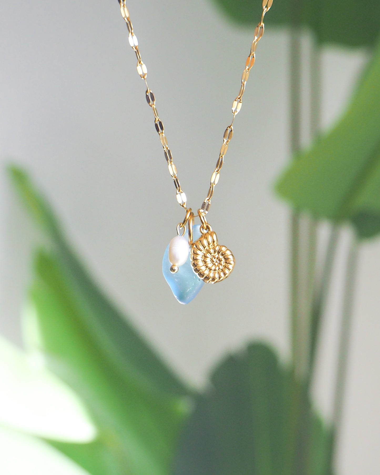 Front view of Oceanic Trio Nautilus Blue Sea Glass Pearl Necklace with green plant background, nautilus shell necklace, ocean blue necklace, coastal jewelry, freshwater pearl necklace, gold seashell necklace, ocean-inspired jewelry, beach necklace, ocean treasure necklace
