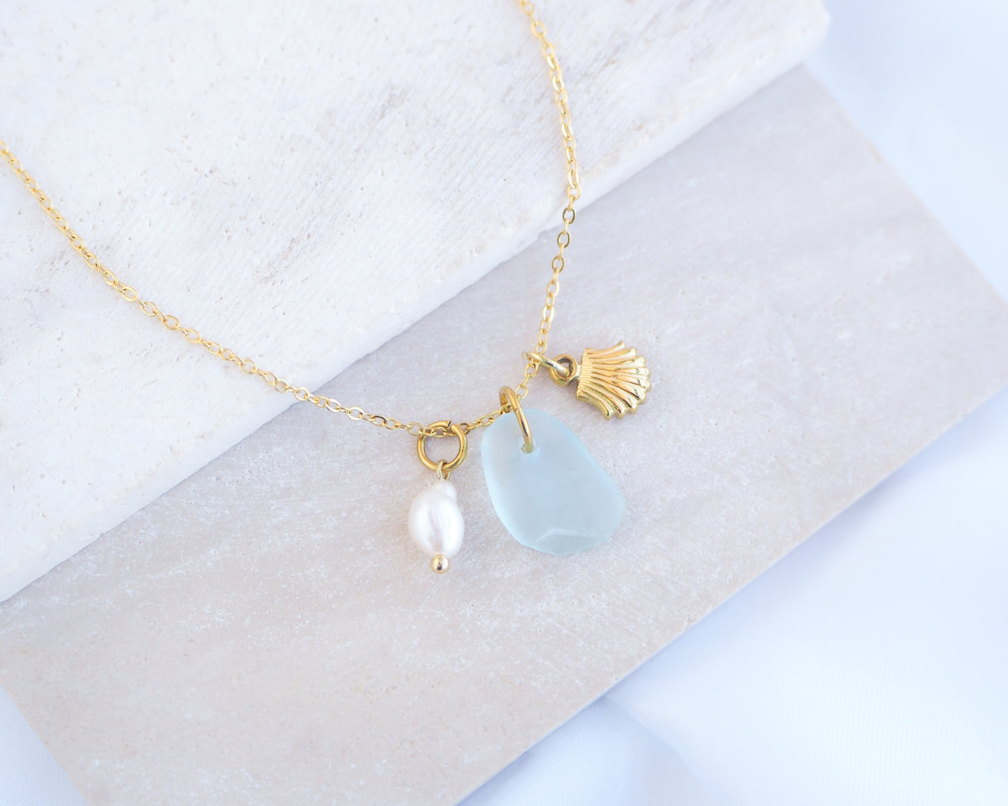 Oceanic Trio Shell Light Blue Sea Glass Pearl Necklace on stone displayed, scallop shell necklace, ocean blue necklace, coastal jewelry, freshwater pearl necklace, gold seashell necklace, ocean-inspired jewelry, beach necklace, ocean treasure necklace