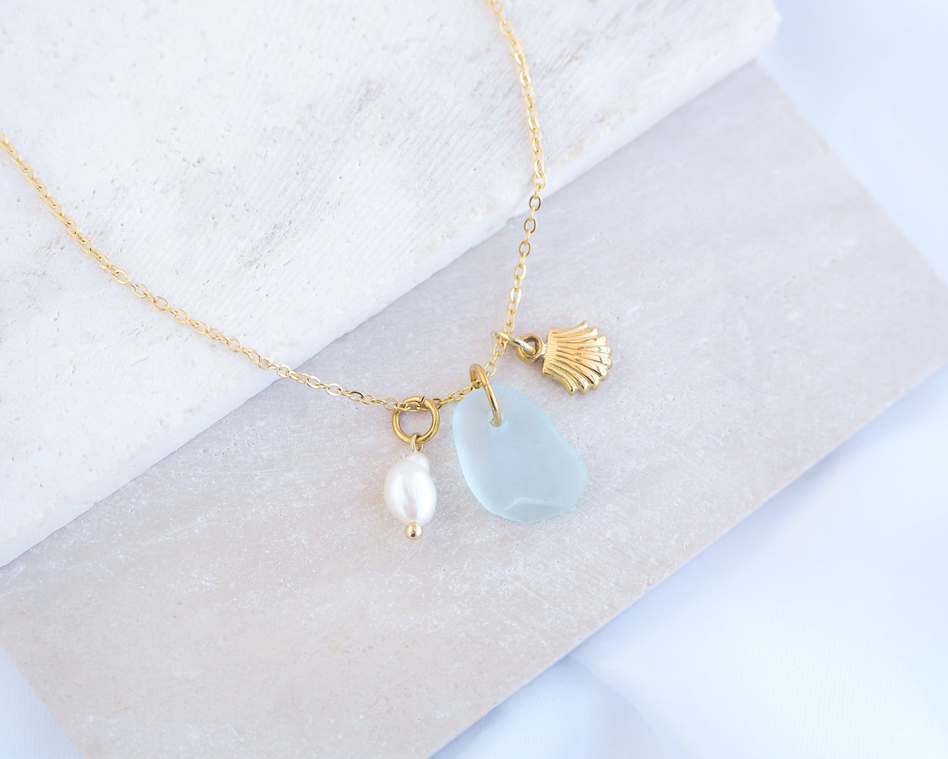Oceanic Trio Shell Light Blue Sea Glass Pearl Necklace on stone displayed, scallop shell necklace, ocean blue necklace, coastal jewelry, freshwater pearl necklace, gold seashell necklace, ocean-inspired jewelry, beach necklace, ocean treasure necklace