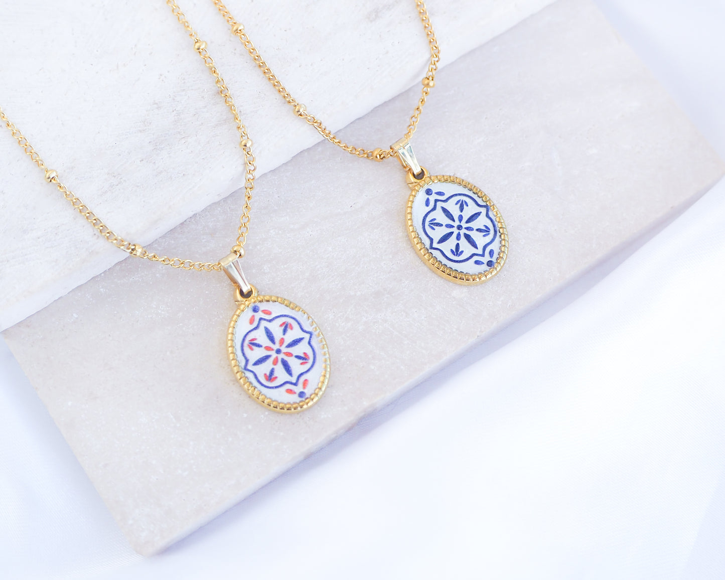 Two gold Portuguese Tile Oval Necklaces on a white stone surface. Made with 18k gold stainless steel and white blue mosaic pendant inspired by the Portuguese Azulejo design pattern. 