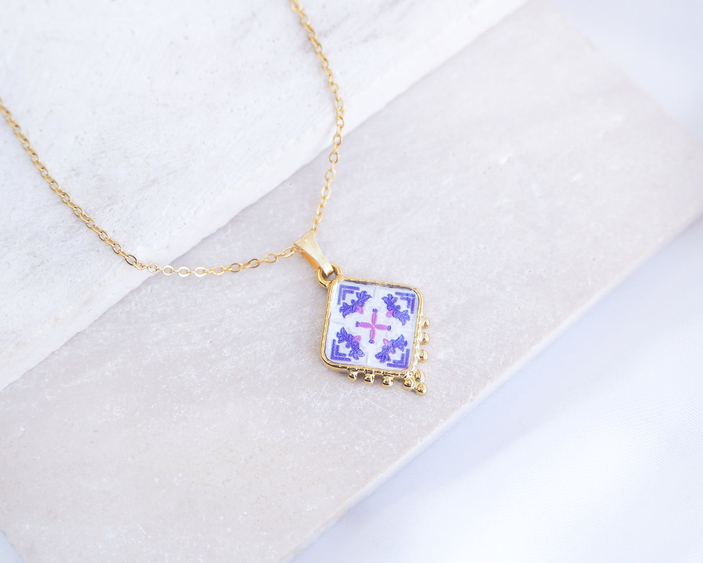 Close up of Portuguese Tile Necklace Gold on stone displayed. Made with 18k goold stainless steel and pink purple pendant inspired by the Portuguese Azulejo design pattern. 