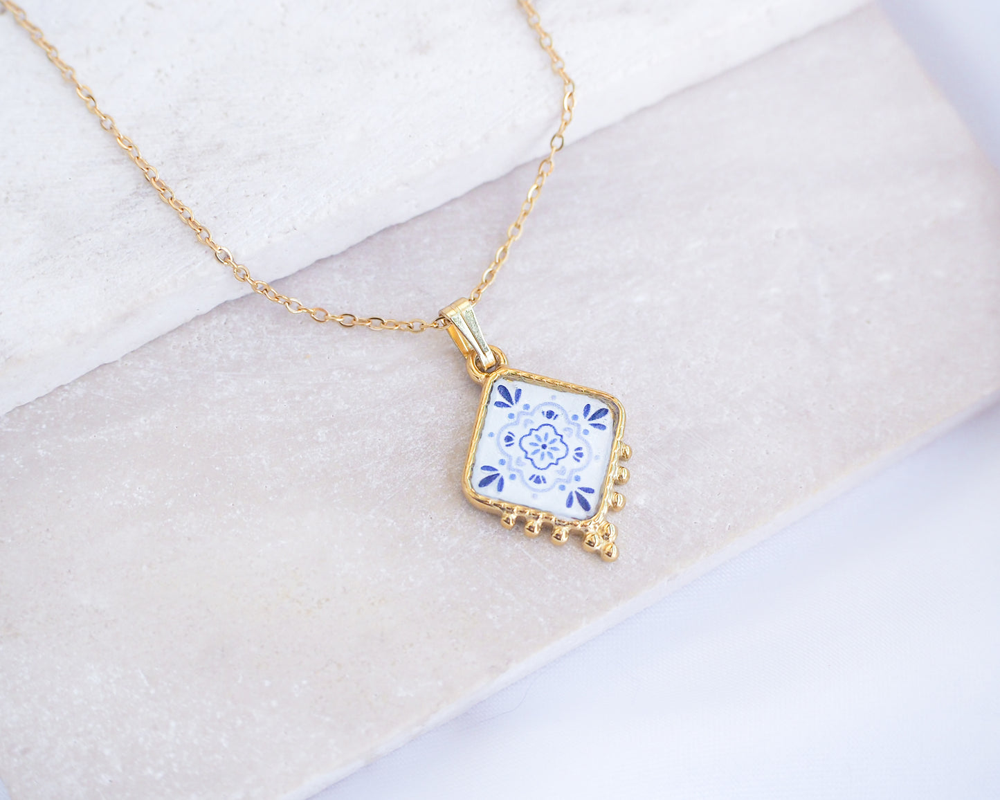 Close up of Portuguese Tile Necklace Blue Blossom Gold on stone displayed. Made with 18k gold stainless steel and white blue floral pendant inspired by the Portuguese Azulejo design pattern. 
