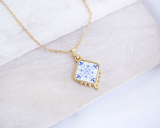 Close up of Portuguese Tile Necklace Blue Blossom Gold on stone displayed. Made with 18k gold stainless steel and white blue floral pendant inspired by the Portuguese Azulejo design pattern. 