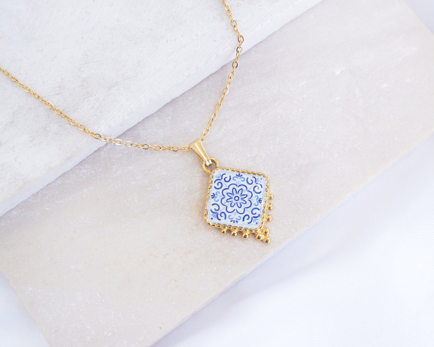 Close up of Portuguese Tile Necklace Floral Blue Gold on stone displayed. Made with 18k gold stainless steel and white blue floral pendant inspired by the Portuguese Azulejo design pattern. 