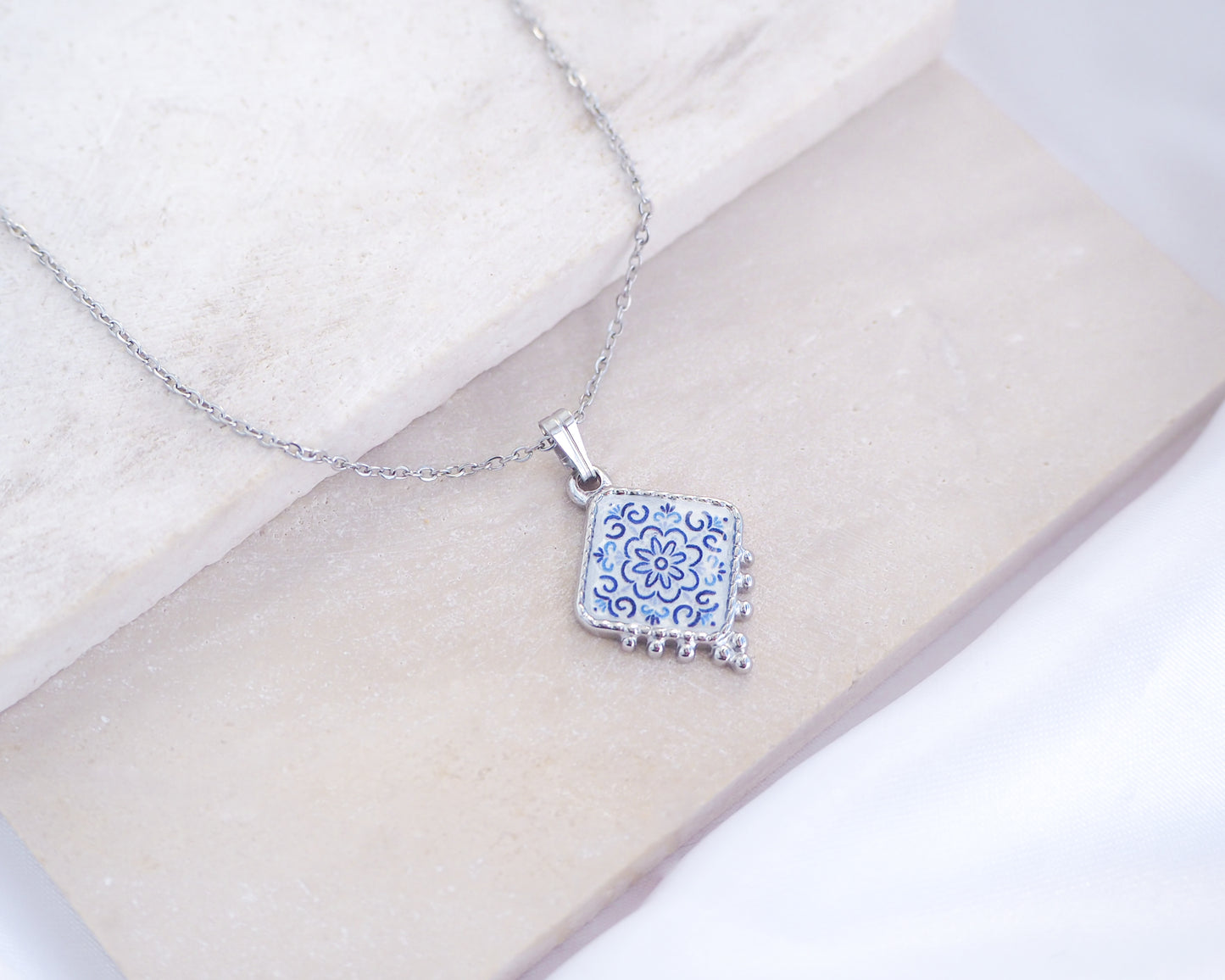 Close up of Portuguese Tile Necklace Floral Blue Silver on stone displayed. Made with stainless steel and white blue floral pendant inspired by the Portuguese Azulejo design pattern. 