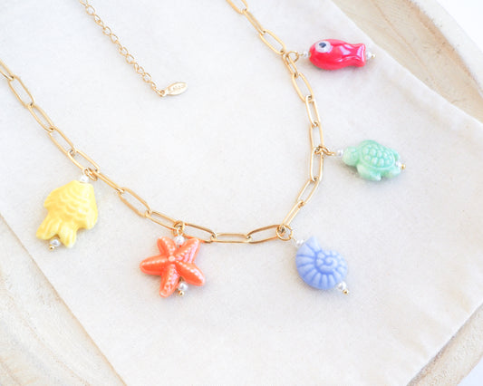 Rainbow Ceramic Charm Necklace with 5 ceramic ocean charms (turtle, starfish, fish, shell & more), sea treasure pendants, 18k gold stainless steel chain displayed on stone 