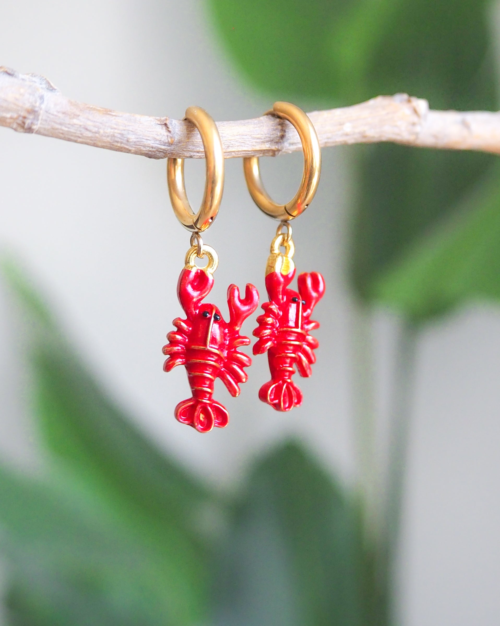 Front view of Red Lobster Gold Earrings with green plant background. Made with 18k gold stainless steel. 