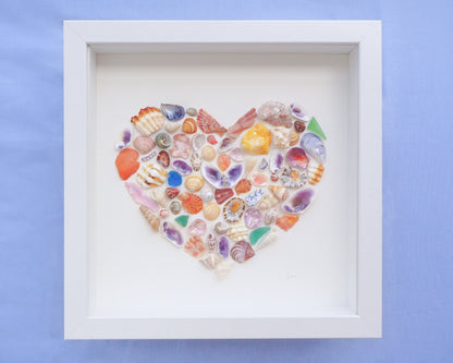 Heart-shaped arrangement of colorful seashells in a white frame on a blue background. Close up of Rainbow Seashell Heart Artwork with colorful shell handpicked from Portugal. Coastal Home Wall Decor, Shell Wall Art, Handmade Shell Artwork, Cute Coastal Nursery Decoration 