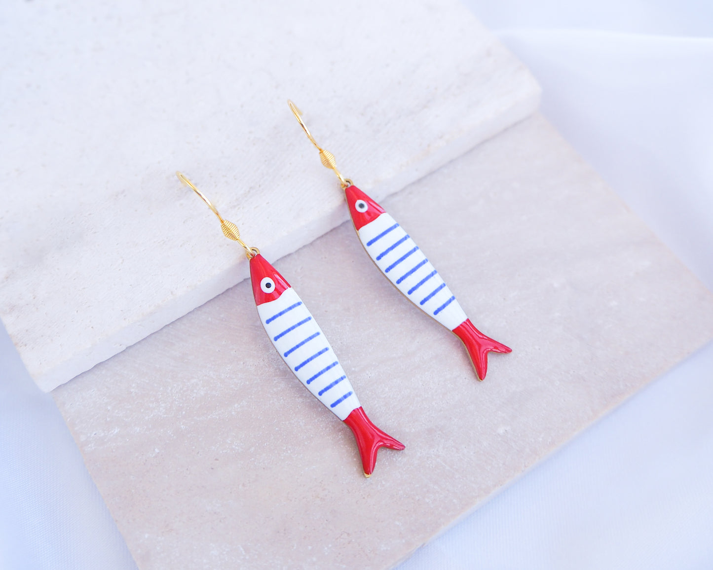 Fish-shaped earrings with red and blue stripes on a light fabric background. Nautical Striped Sardine Fish Earrings with 14k gold stainless steel earring hooks displayed on stone, Red Blue White Striped Fish Pendant, Beautiful gift for Portugal lovers or holiday memory, coastal jewelry from Portugal