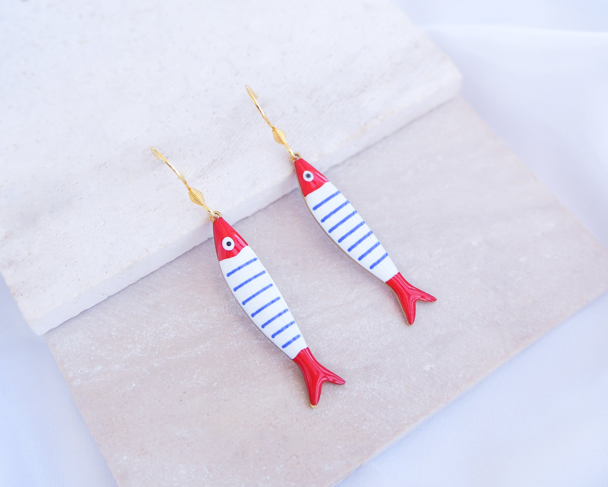 Fish-shaped earrings with red and blue stripes on a light fabric background. Nautical Striped Sardine Fish Earrings with 14k gold stainless steel earring hooks displayed on stone, Red Blue White Striped Fish Pendant, Beautiful gift for Portugal lovers or holiday memory, coastal jewelry from Portugal
