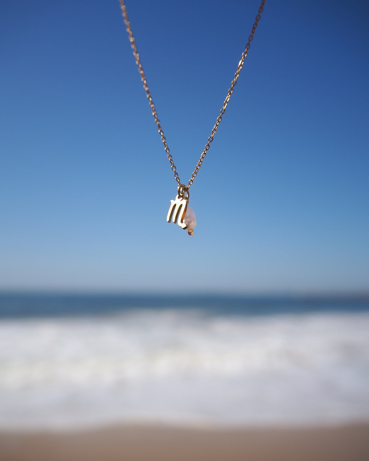 Scorpio pearl pendant necklace against a blue sky and ocean background. Made of 18k gold stainless steel and a freshwater pearl. The pendant is showing a scorpio zodiac sign charm. 