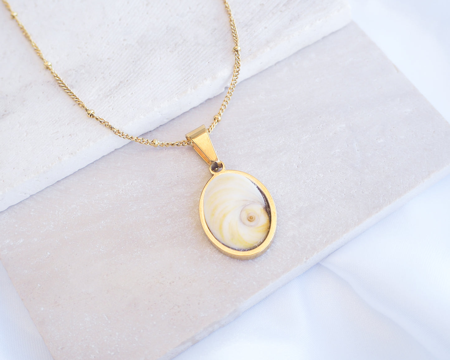 Light Shiva Eye Necklace II on stone displayed. Light Eye of Saint handpicked from Portugal, 18k gold stainless steel shiva eye pendant necklace, Saint Lucy Eye, operculum pendant
