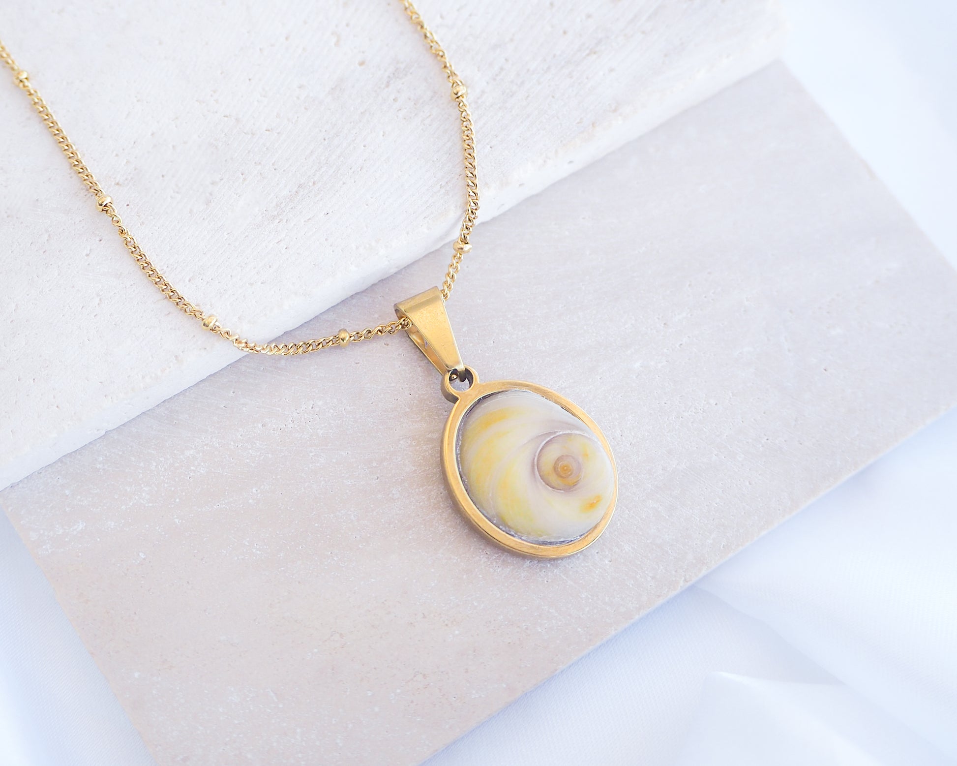 Soft Yellow Shiva Eye Necklace III on stone displayed. Light Eye of Saint handpicked from Portugal, 18k gold stainless steel shiva eye pendant necklace, Saint Lucy Eye, operculum pendant