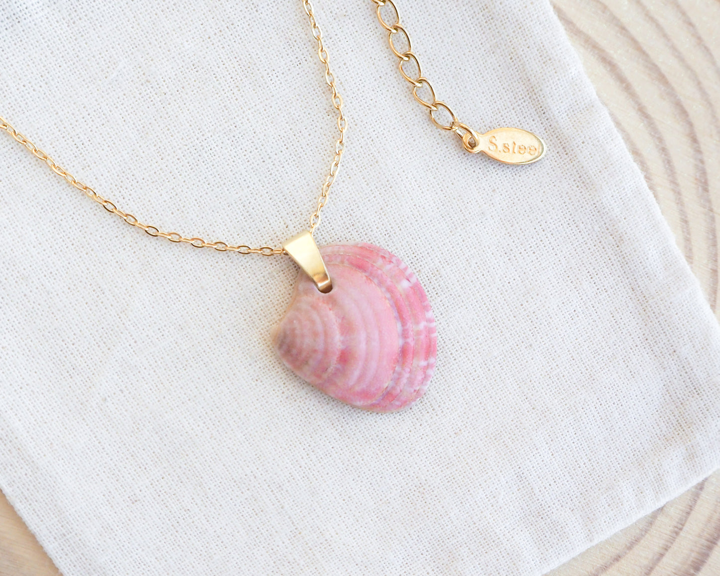 Necklace with a pink shell pendant on a white fabric background. Gold Pink Venus Shell Necklace on cotton displayed. Pink Shell from Portugal. Necklace made with 18k gold stainless steel