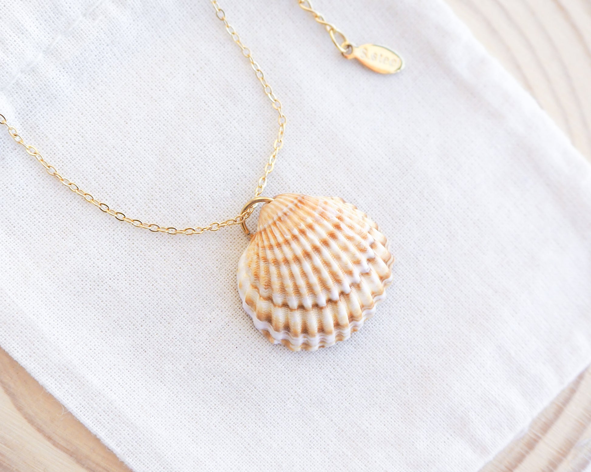A gold cockle shell necklace made with a Mediterranean Cockle Shell from Portugal on a 18k gold stainless steel chain against a cotton background. The shell is yellow with white stripes.