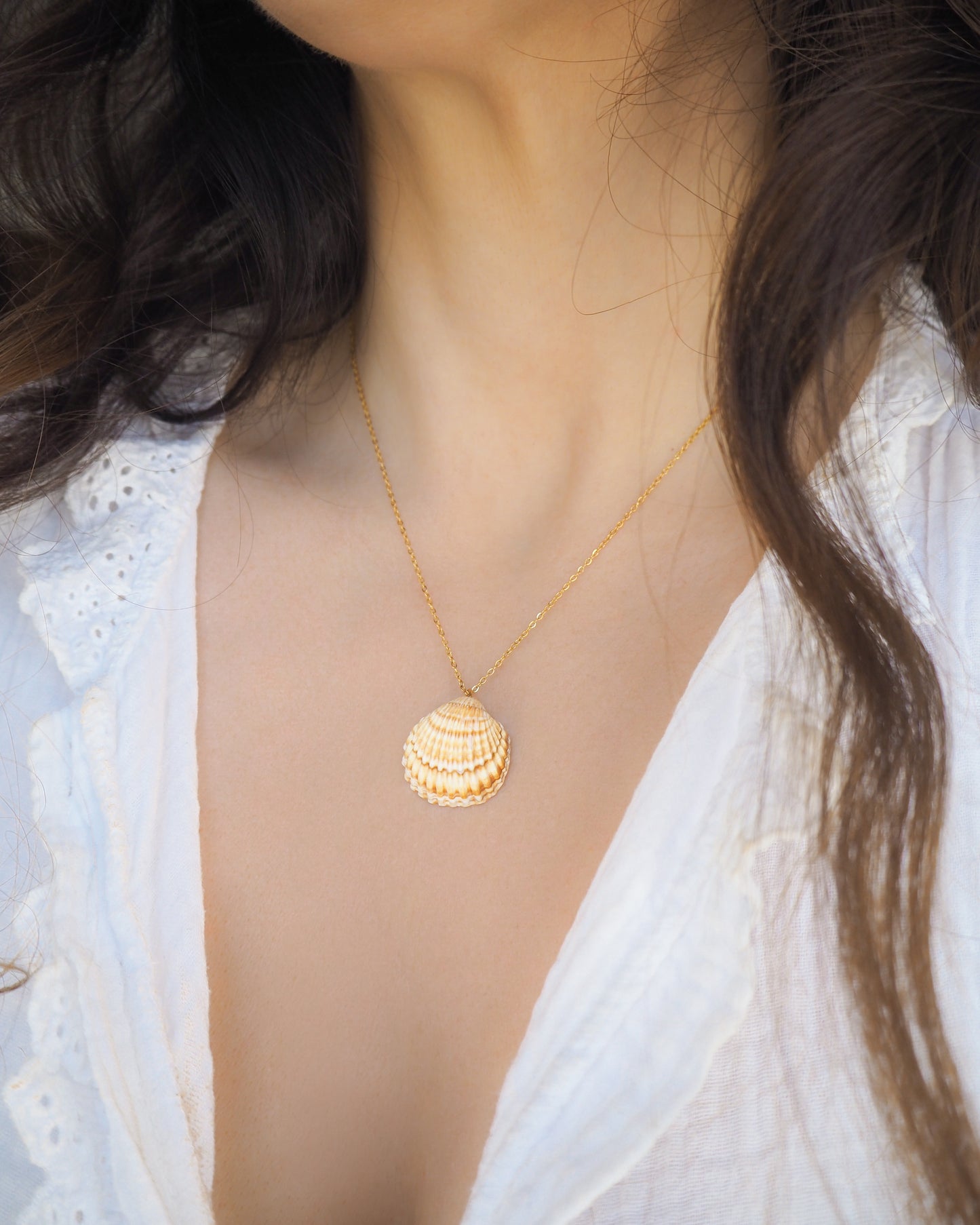 Model wearing a gold cockle shell necklace on neck with white shirt. The necklace is made with a Mediterranean Cockle Shell from Portugal on a 18k gold stainless steel chain The shell is yellow with white stripes.