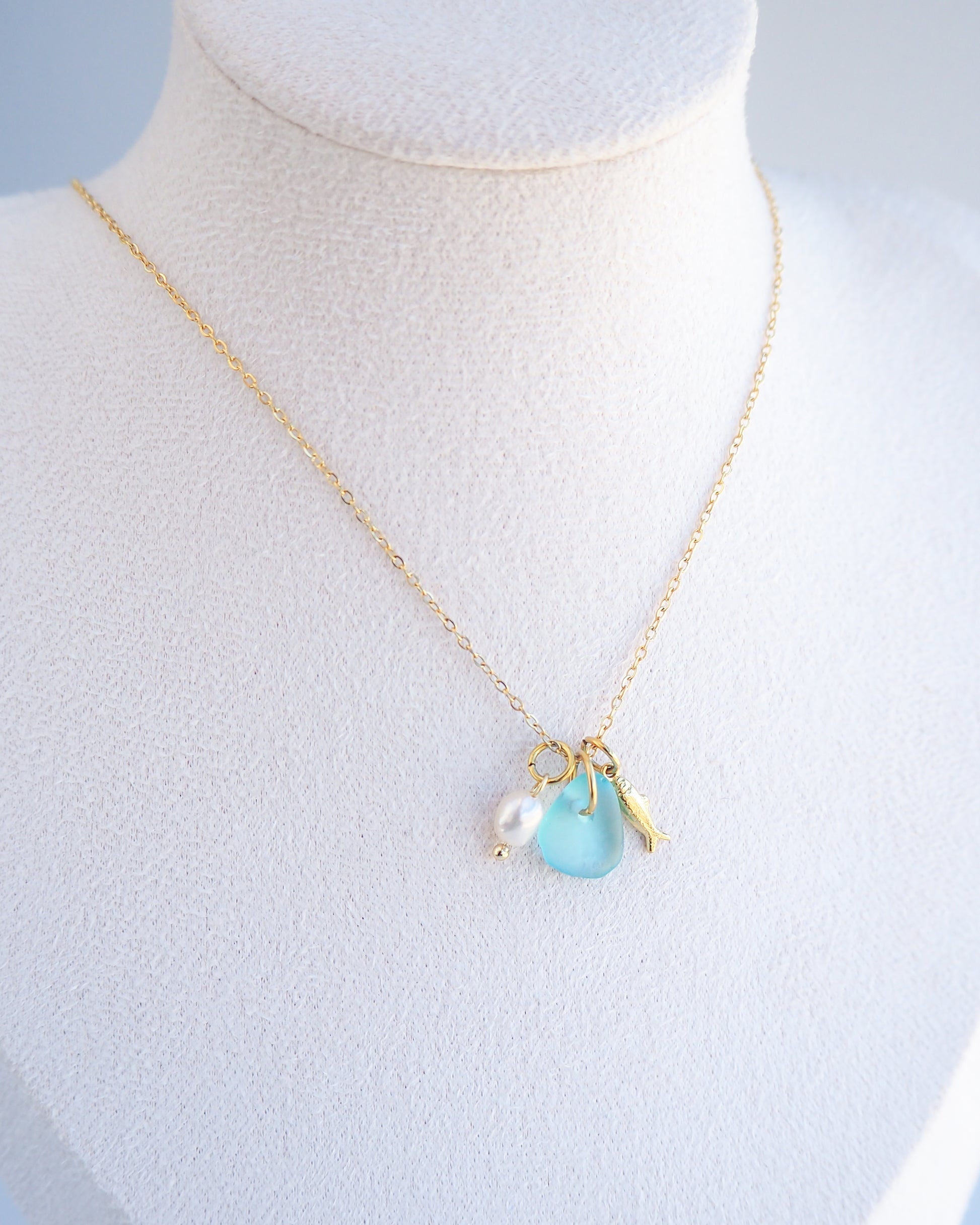 Close up of a necklace with a tiny sardine fish pendant, turquoise blue sea glass and freshwater pearl pendant on display. Made with gold stainless steel chain.