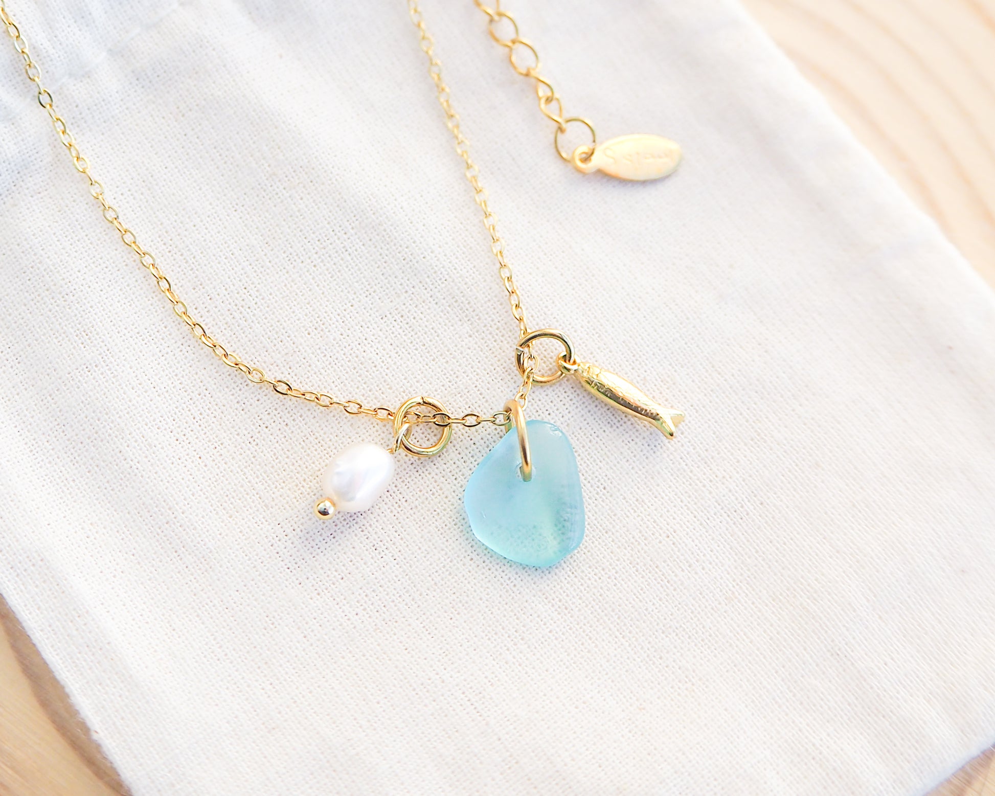 Close up of a necklace with a tiny sardine fish pendant, turquoise blue sea glass and freshwater pearl pendant on cotton fabric. Made with gold stainless steel chain.