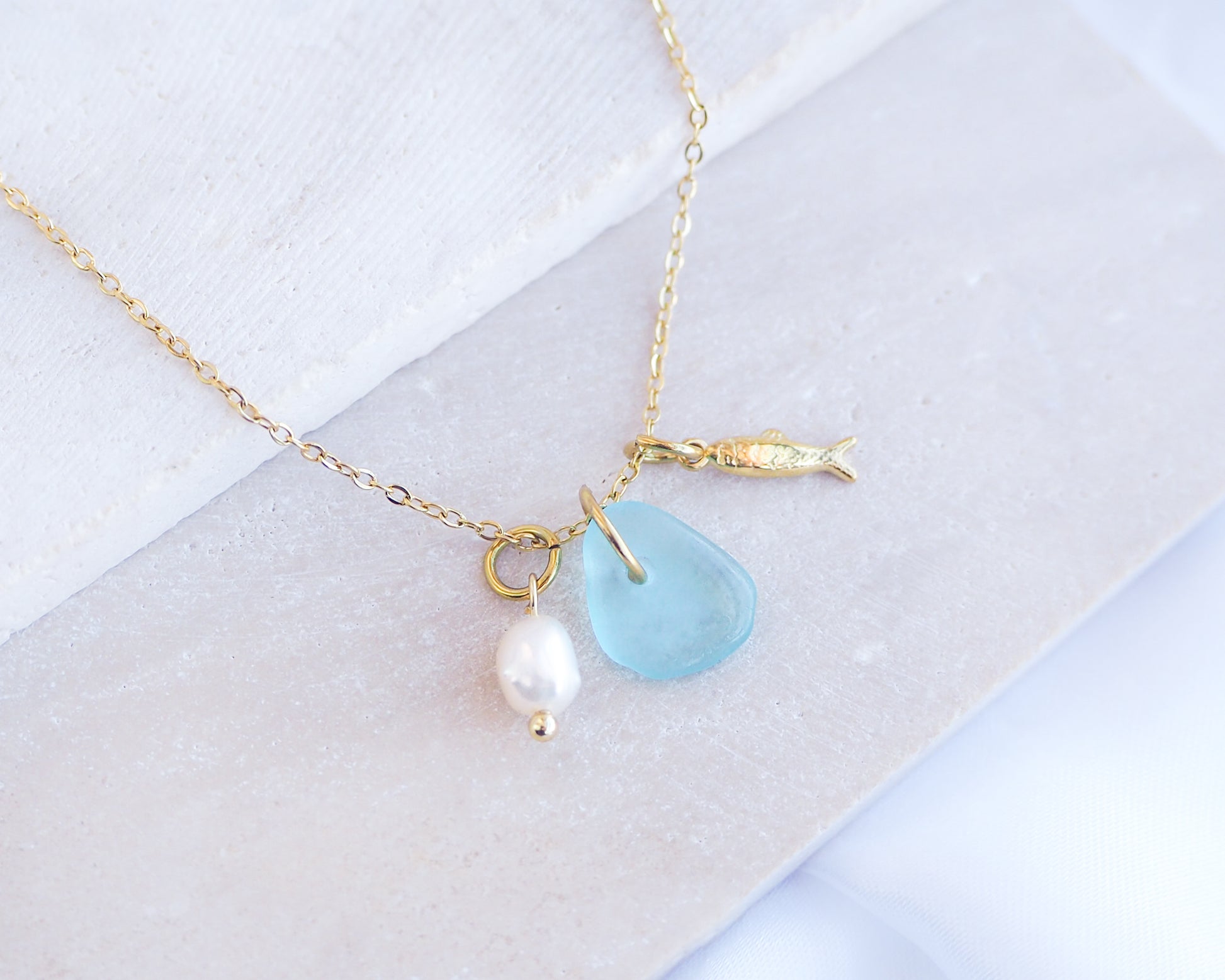 Close up of a necklace with a tiny sardine fish pendant, turquoise blue sea glass and freshwater pearl pendant on a white stone background. Made with gold stainless steel chain.
